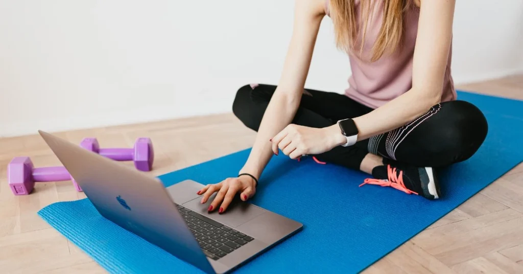 Fitness student sitting on a gym mat with a laptop and dumbbells, studying for a personal training course